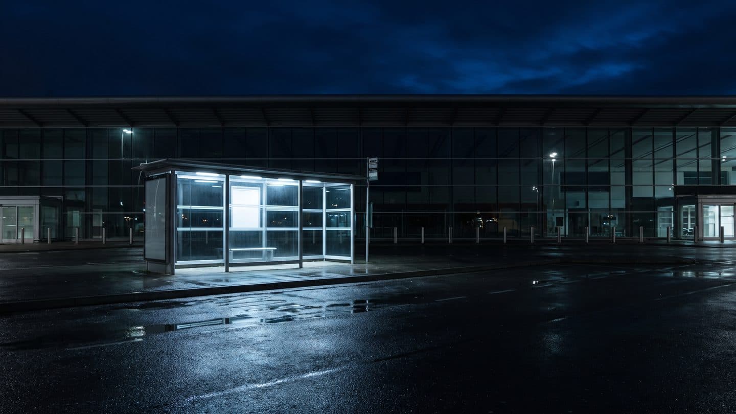 Pusta szklana wiata przystankowa podświetlona od wewnątrz, stojąca nocą na opustoszałym podjeździe przed lotniskiem, mokry asfalt odbija światło lamp, ani pojazdów, ani ludzi
