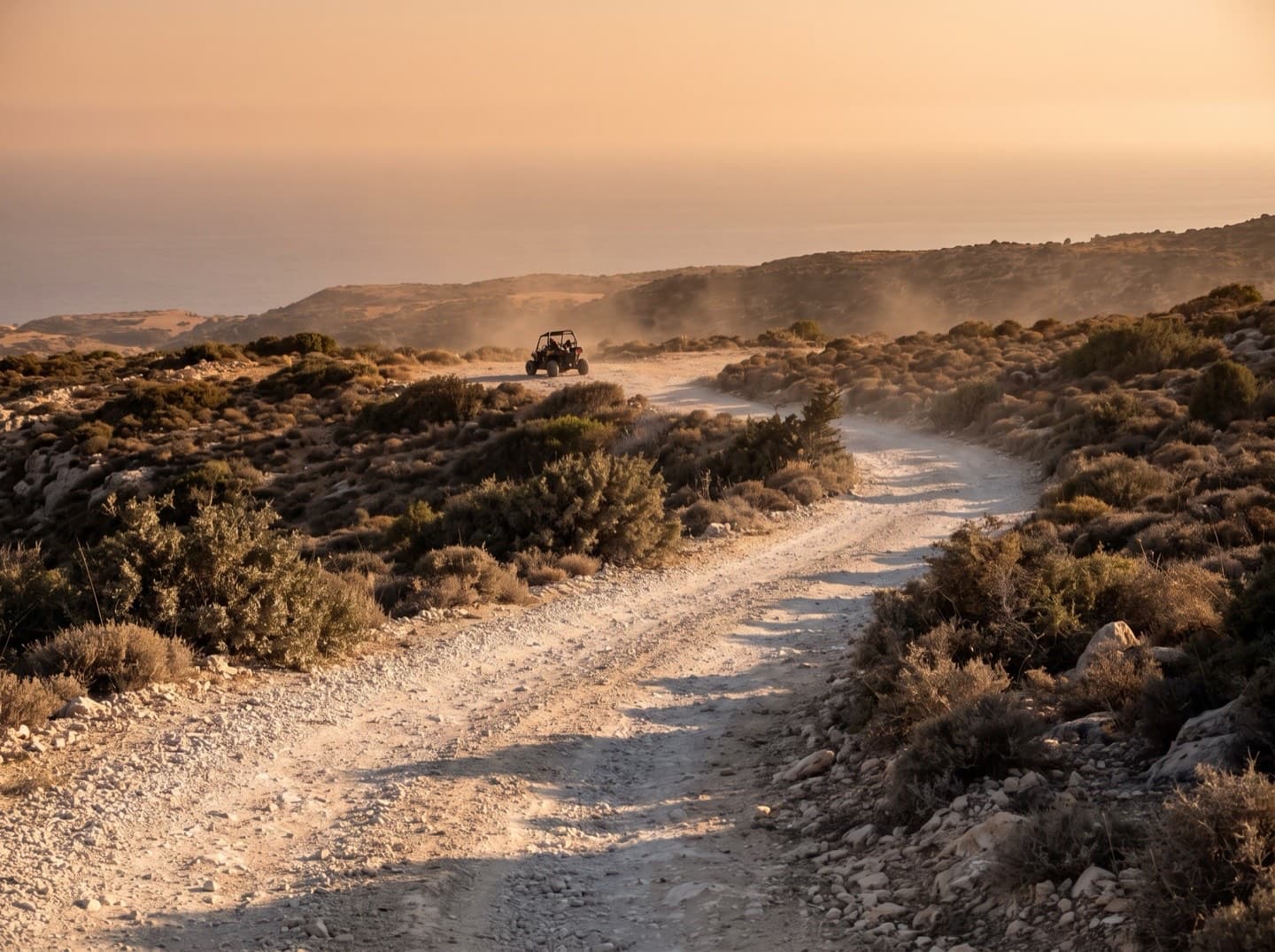 Eine steinige Schotterpiste, die sich durch niedrige Macchia an einem Kalksteinhang zu einem dunstigen Meereshorizont zieht, weit hinten ein kleiner Offroad-Buggy
