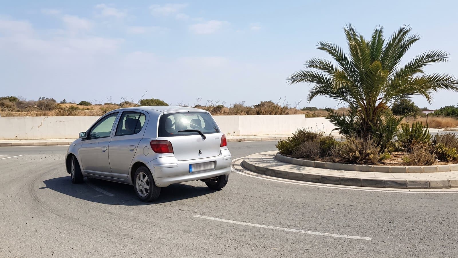 The view from behind a small hatchback bearing left into a Cyprus roundabout, the planted island on its right