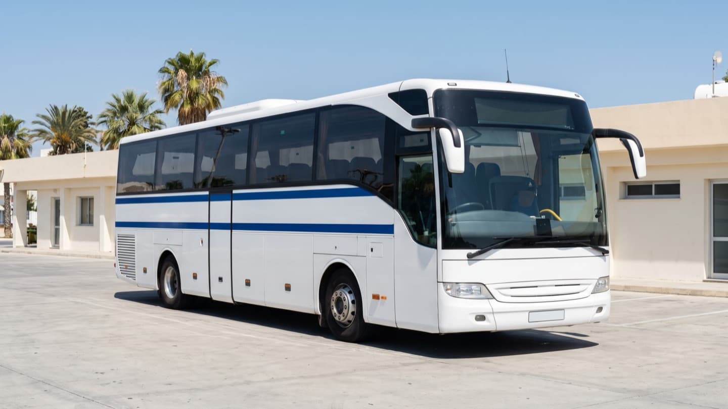 A modern intercity coach at a kerbside stop on a wide city avenue in bright daylight, a few passengers boarding with luggage, palm trees and low apartment buildings behind
