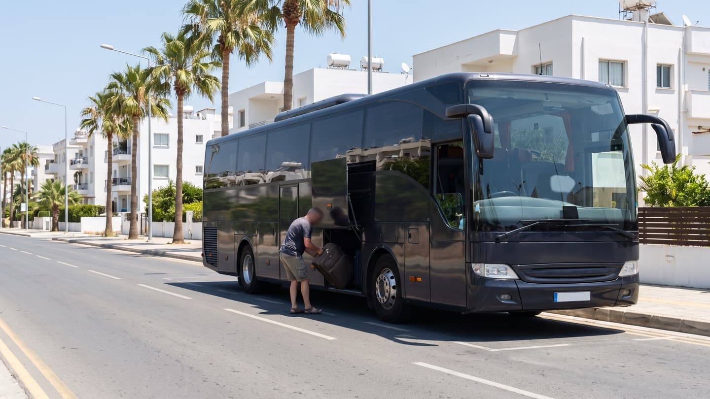 A modern intercity coach at a kerbside stop on a wide avenue lined with palms and low white buildings, a passenger loading a suitcase into the luggage bay