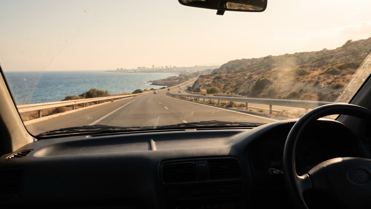 Driver's-eye view through a windscreen of a right-hand-drive car on an open coastal motorway, sea to the left, dry hills to the right, a distant city on the horizon